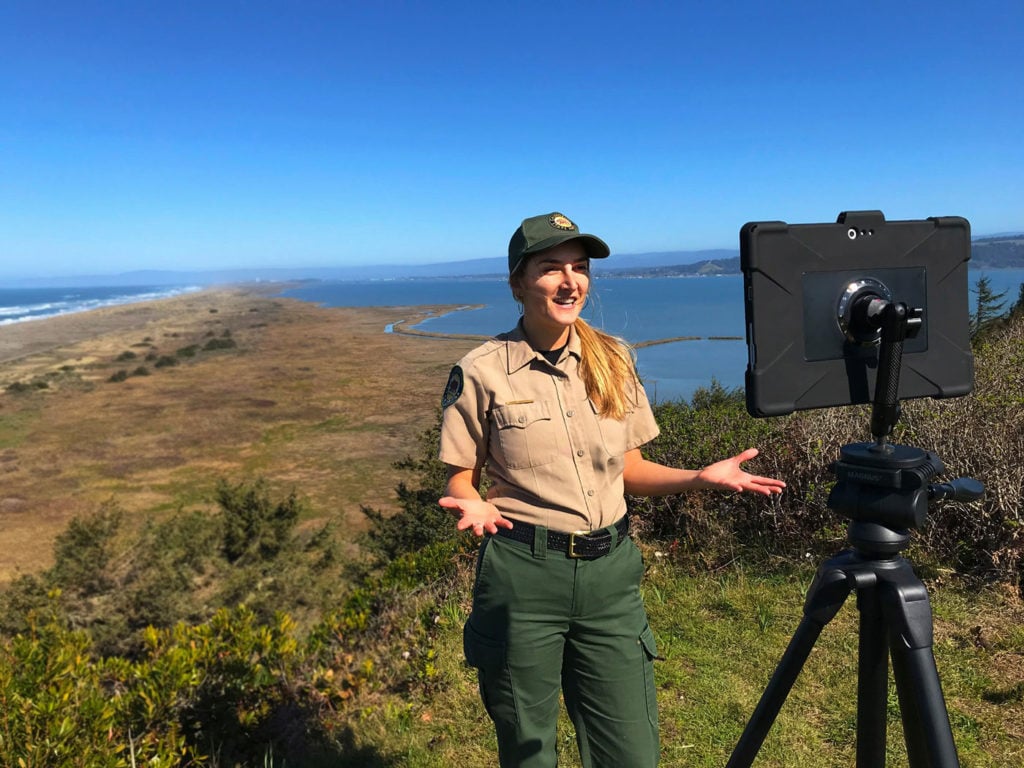 A female California State Parks naturalist interpreter in park ranger fatigues gives her presentation before a tablet on a tripod on which she is livestreaming a virtual program to Facebook.