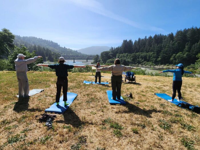 Several people stand on yoga mats on a sunny day, looking out across a grassy field to a river and redwood-lined hillsides