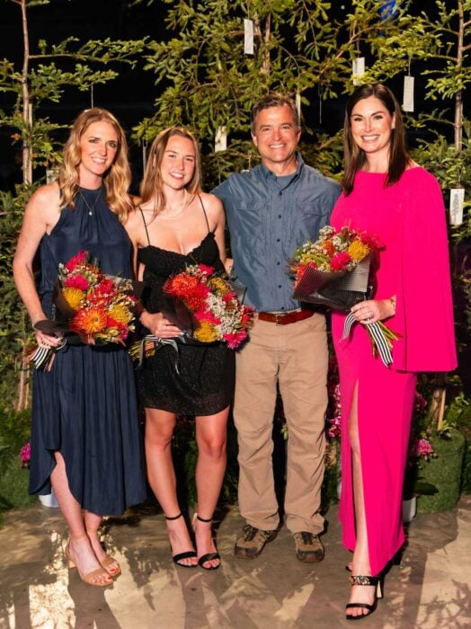 Three women in evening wear holding bouquets stand with a man, all of them smiling for the camera