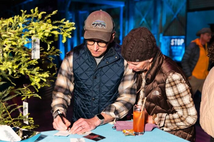 Two people wearing flannel write on pieces of paper next to a wish tree.